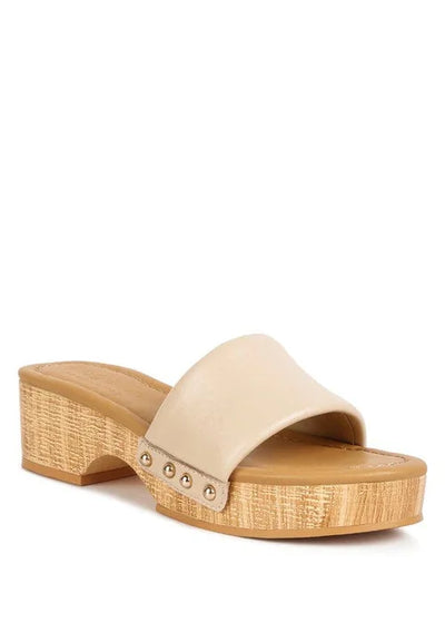Beige slide sandal with wooden platform heel and metal stud detail on white background