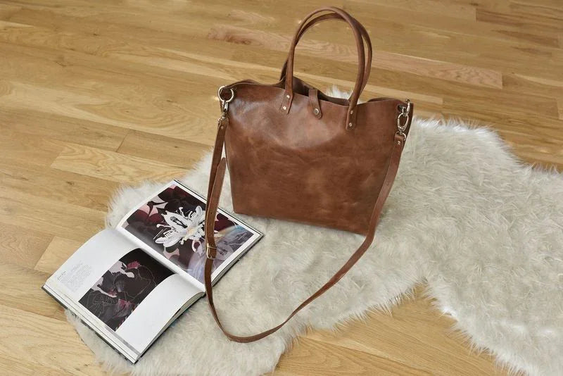Brown leather tote bag with shoulder strap on white rug beside open magazine