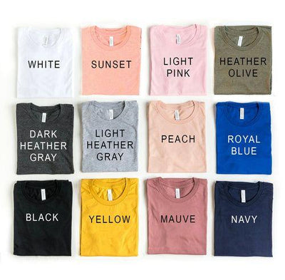 Twelve folded t-shirts in different colors labeled white, sunset, light pink, heather olive, dark heather gray, light heather gray, peach, royal blue, black, yellow, mauve, navy.