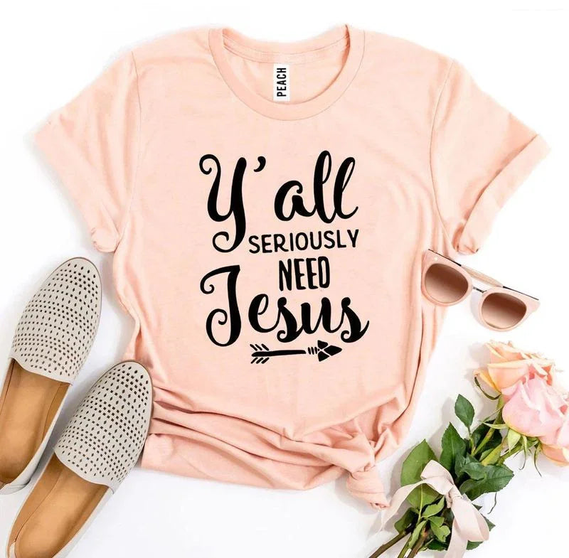 Peach t-shirt with 'Y'all seriously need Jesus' text, beside beige shoes, sunglasses, pink roses.