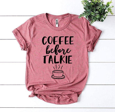Pink graphic t-shirt with 'Coffee Before Talkie' slogan and coffee cup, flat lay with plant.