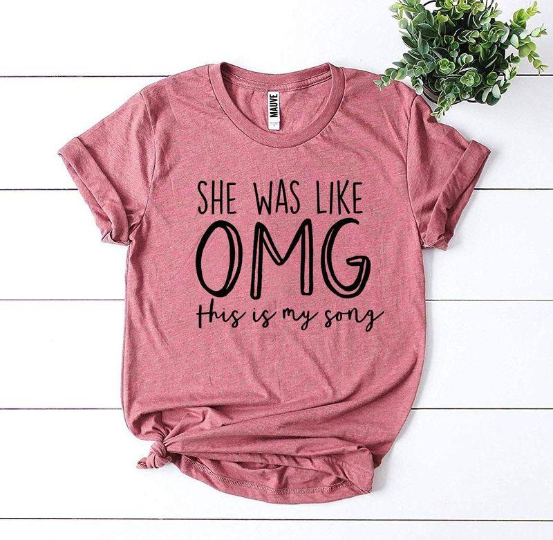 Pink graphic tee with 'She was like OMG this is my song' text, white wood background, plant