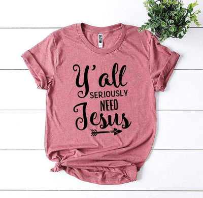 Pink graphic tee with 'Y'all seriously need Jesus' text, flat lay on white wooden background.