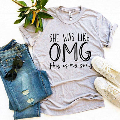 Flat lay of gray graphic t-shirt with 'She was like OMG this is my song', jeans, shoes, sunglasses.