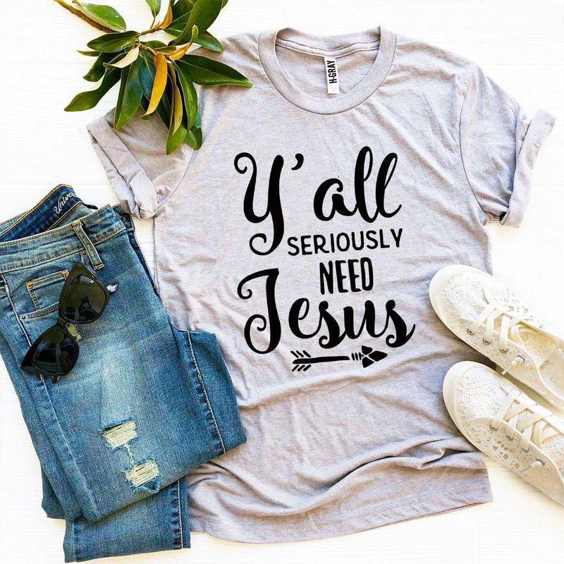 Gray graphic tee with 'Y'all Seriously Need Jesus' text, styled with jeans, sneakers, sunglasses, and a plant.
