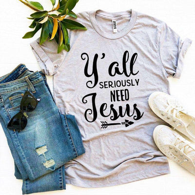 Gray graphic tee with 'Y'all Seriously Need Jesus' text, styled with jeans, sneakers, sunglasses, and a plant.