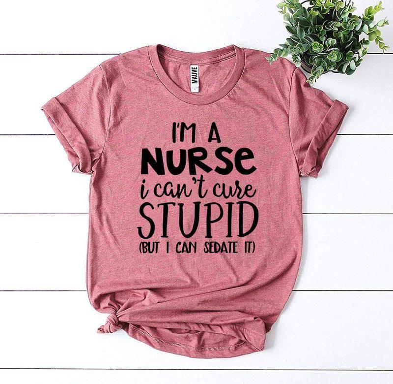 Pink nurse humor t-shirt with 'I'm a nurse, I can't cure stupid' text on white wood background