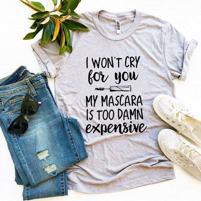 Flat lay of a gray graphic t-shirt with 'I won't cry for you my mascara is too damn expensive' text, denim jeans, lace sneakers, black sunglasses, and a plant, casual fashion outfit.